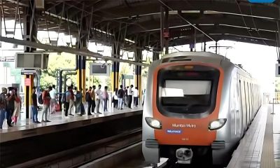 Mumbai Metro train arriving at a station with passengers waiting on the platform. The train features an orange and silver design. The station is covered with a metal roof, and digital signboards are visible above the platform.