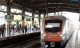 Mumbai Metro train arriving at a station with passengers waiting on the platform. The train features an orange and silver design. The station is covered with a metal roof, and digital signboards are visible above the platform.