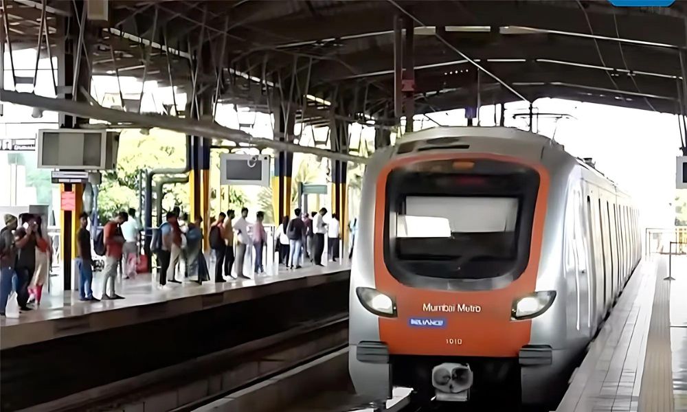 Mumbai Metro train arriving at a station with passengers waiting on the platform. The train features an orange and silver design. The station is covered with a metal roof, and digital signboards are visible above the platform.