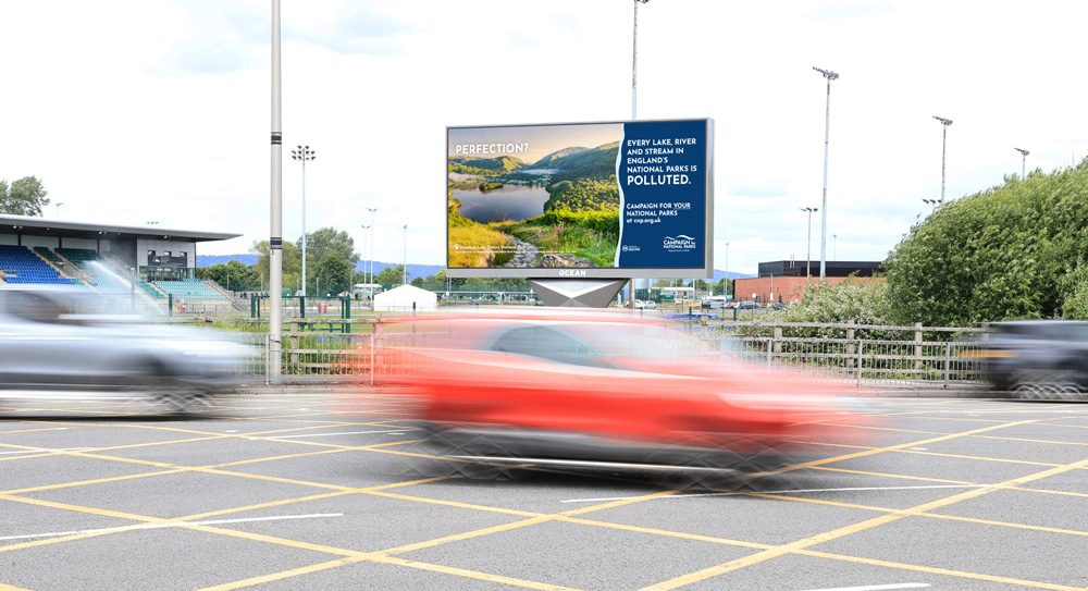 Digital billboard by the roadside promoting the "Campaign for National Parks," highlighting pollution in England's national parks. Blurred cars pass by in the foreground