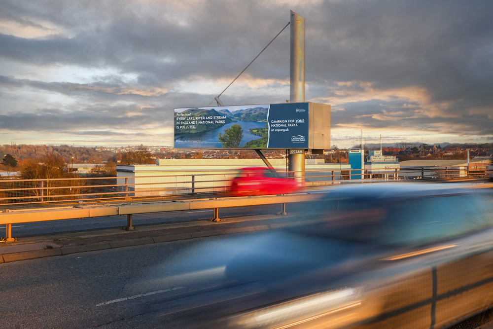 Digital billboard by the roadside promoting the "Campaign for National Parks," highlighting pollution in England's national parks. Blurred cars pass by in the foreground