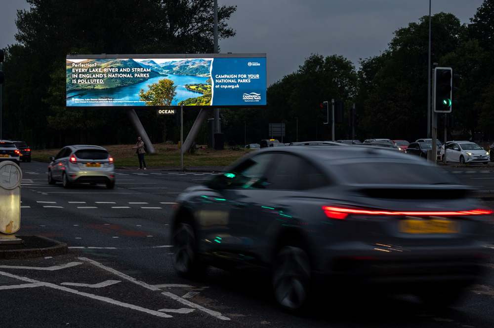 Digital billboard at a busy intersection promoting the "Campaign for National Parks" with cars driving by in low light.